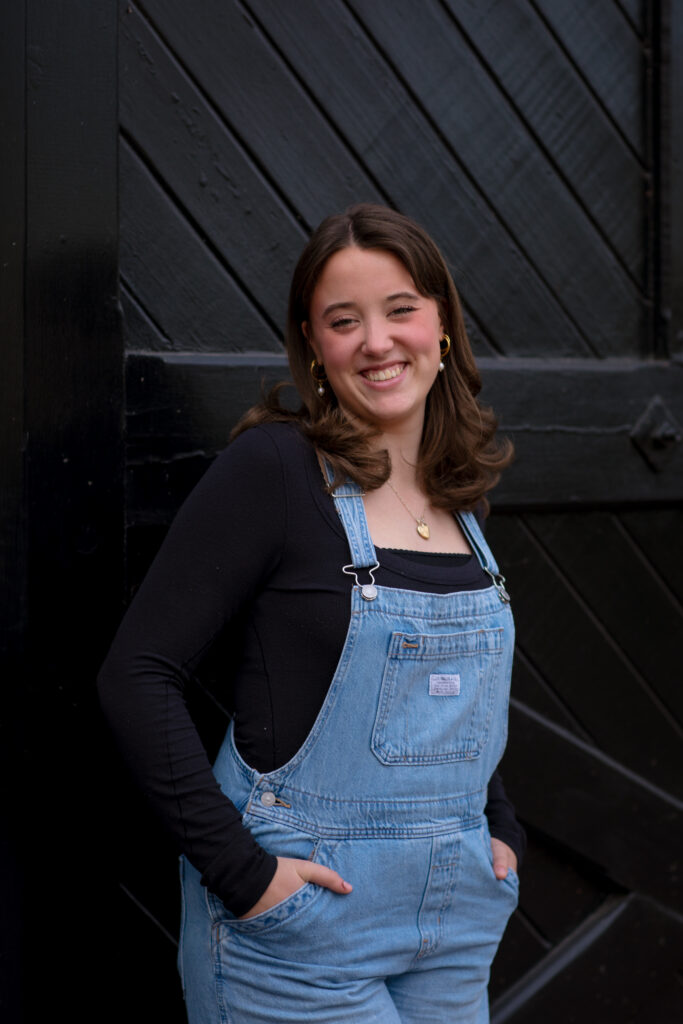 Young woman smiles in front of black barn door with overalls for a Kat Jones Creative headshot